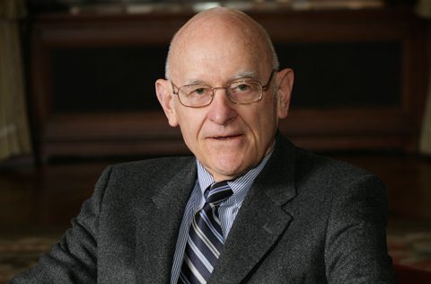 Portrait of George A. Nicholson III, an older gentleman with glasses, wearing a dark suit and striped tie, seated in a red chair with a warm and composed expression. The setting is elegant and professional, reflecting his distinguished career and legacy of service.